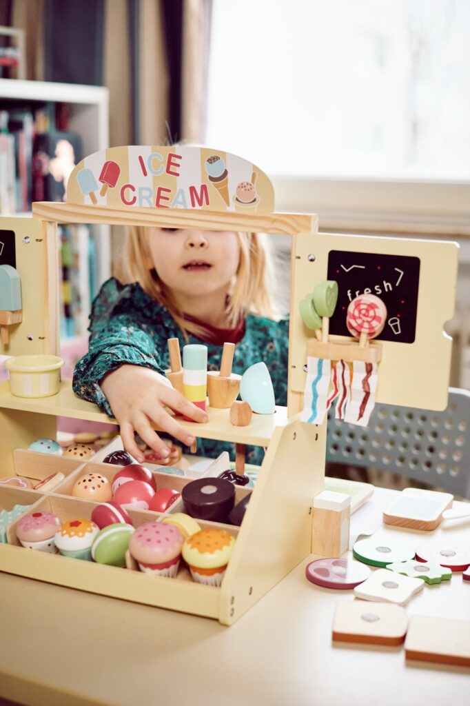 Cute little blond hair girl playing ice cream stand at home. Social skills development in play